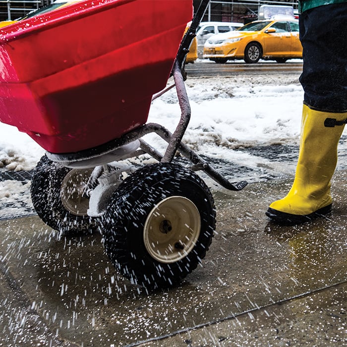 Person wearing yellow boots and pushing a spreading machine full of solid particle ice melt